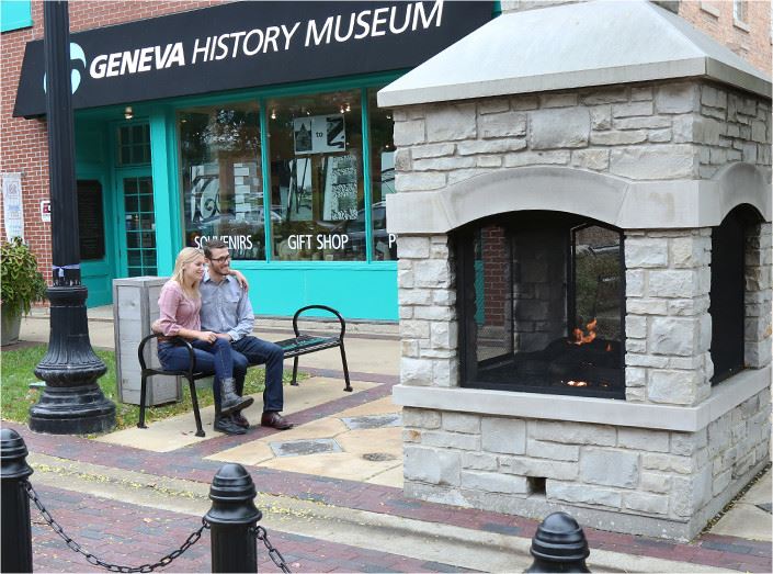 A couple sitting next to an outdoor fireplace with a Geneva-themed gift shop in the background